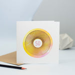 A 50th birthday card featuring a gorgeous pink and yellow watercolour background and a gold foiled spirograph on top with the number 50 in the centre in pink