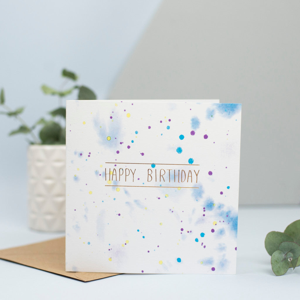 A watercolour splattered background with gold foiled Happy birthday on top
