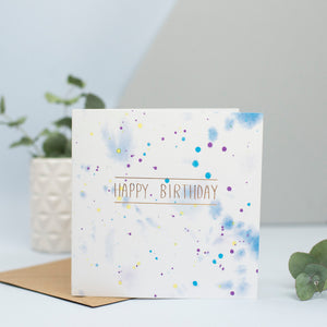 A watercolour splattered background with gold foiled Happy birthday on top