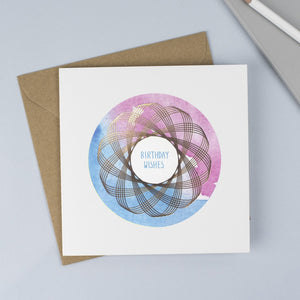 Copper foiled Spirograph with a pink & blue watercolour background and hand drawn font with the words Birthday Wishes in blue.