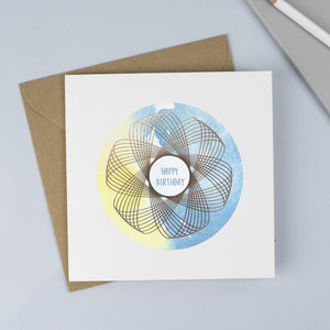 A unisex birthday card with a blue and yellow watercolour background and a gold spirograph on top.